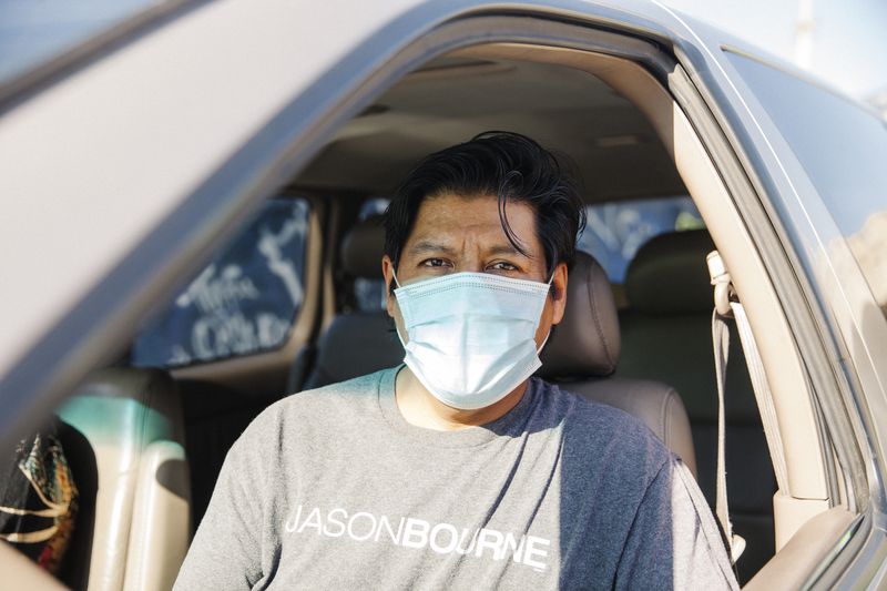 Man wearing disposable mask looks directly at the camera from the front seat of his car.