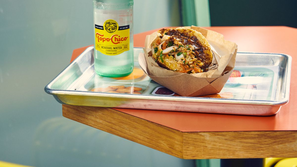 A mushroom Sloppy Joe is shot from the side; it sits on a metal lunch tray on an orange table; a bottle of Topo Chico stands next to the sandwich