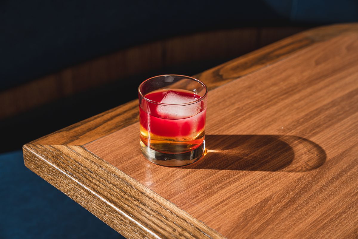 A red and clear cocktail with a large cube of ice sits on the edge of a wooden table.
