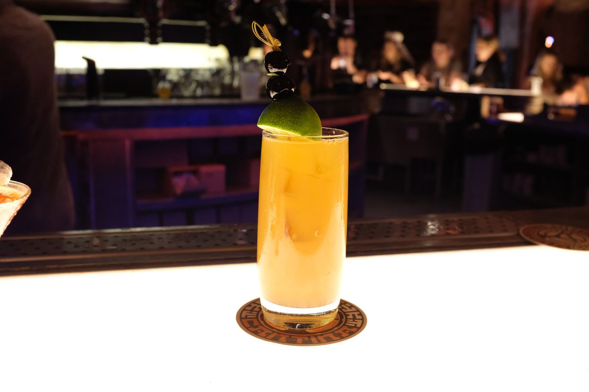 A highball glass with a lime wedge full of an orange cocktail on a coaster on a bright, backlit bar with customers blurred at the bar beyond