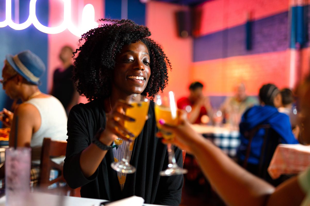 A young Black woman smiles while cheersing a friend with a mimosa at the Breakfast Boys in College Park, GA.