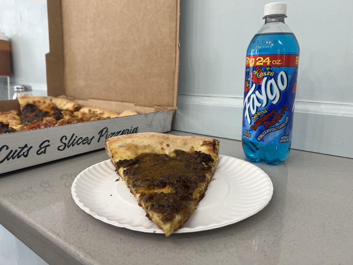 A forest green slice of curried oxtail pizza sits on a paper plate next to a blue raspberry soda