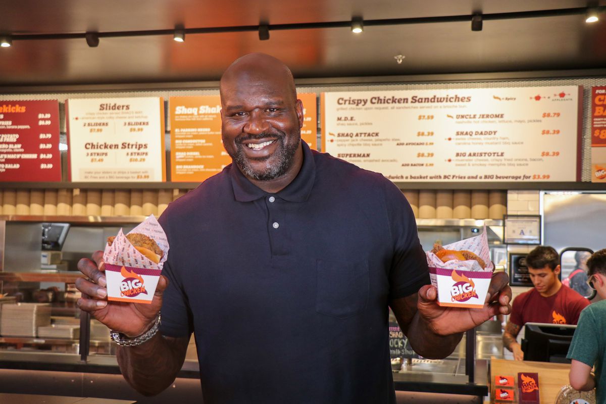 A smiling Shaquille O’Neal holding up a fried chicken sandwich in each hand.