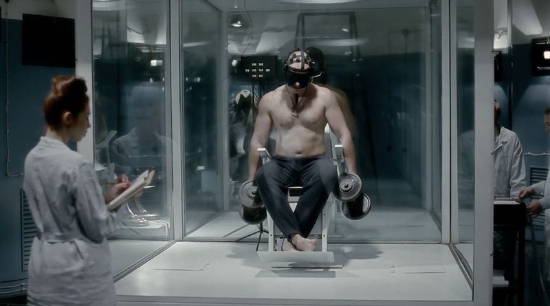 A seated, shirtless, shoeless, masked man in a glass box lifts weights while a uniformed woman observes and takes notes.