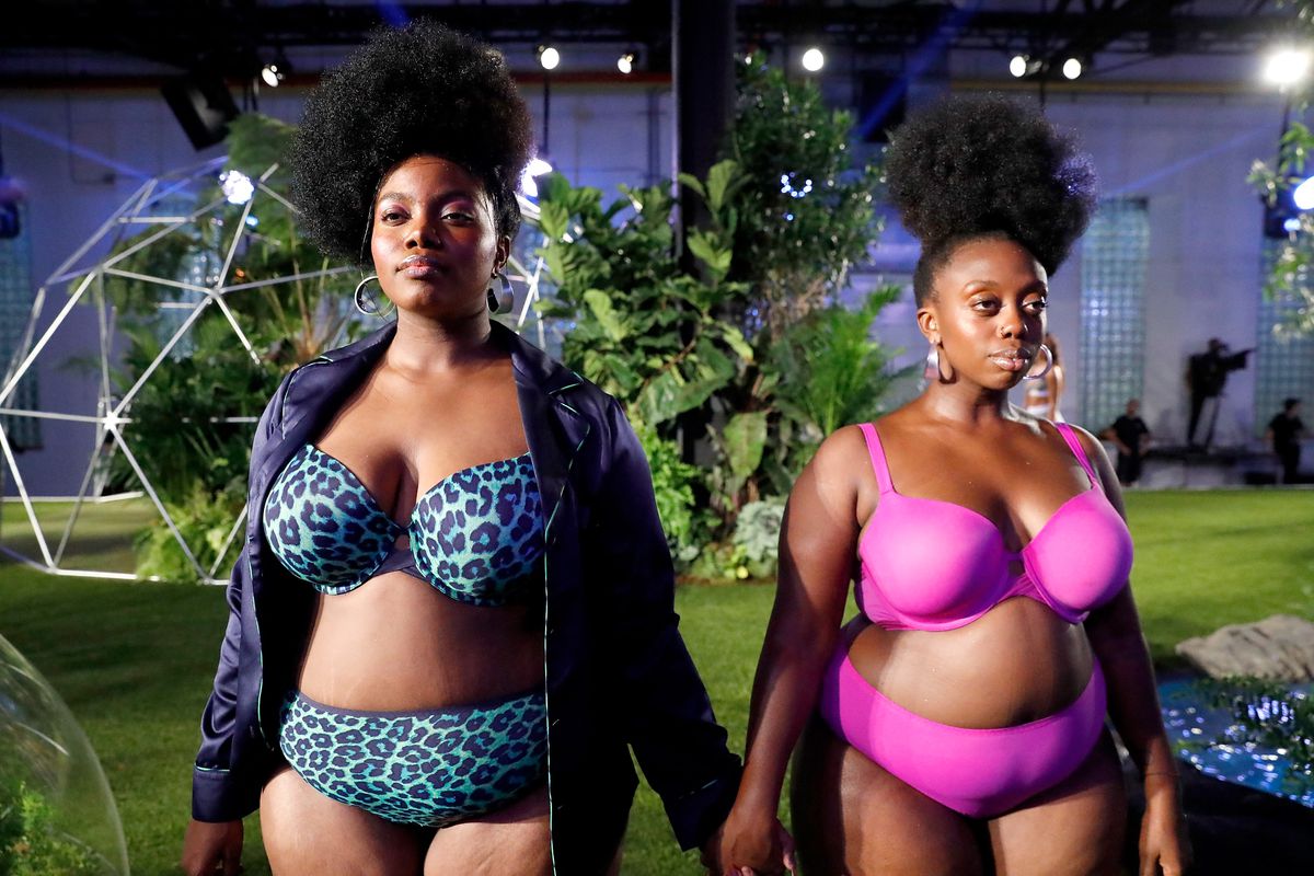 Two models, one wearing a hot pink bra and underwear and the other wearing blue leopard print, stand hand in hand.