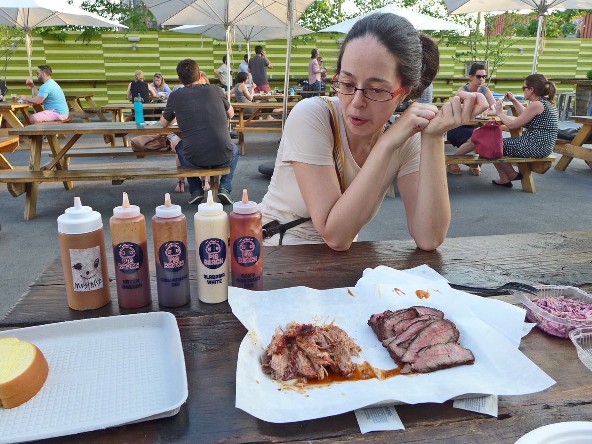 A women sits at a picnic table with two kinds of meat and a thick slice of bread, with tables of diners seen behind her.