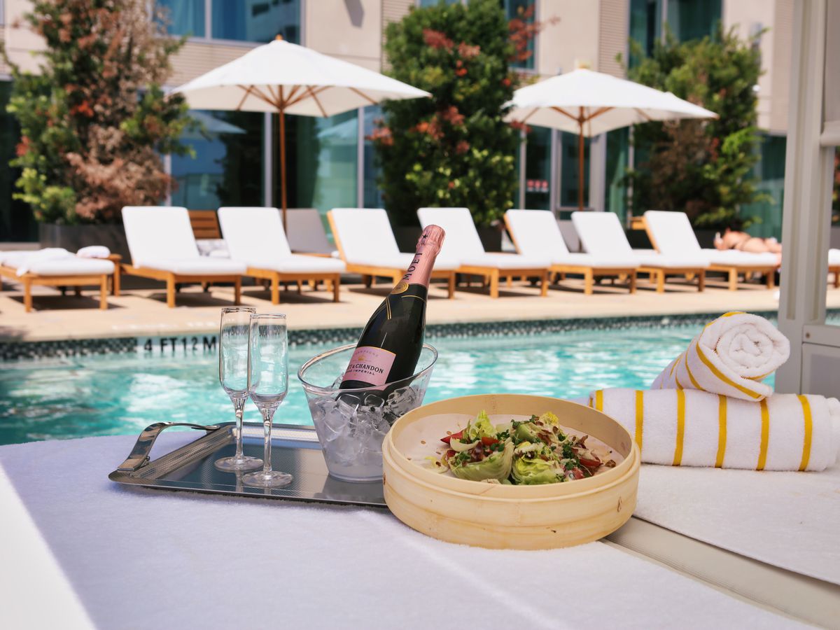 A tray with champagne flutes and a bottle of champagne, rolled towels, and a bamboo tray featuring a salad nearby the Laura Hotel pol.