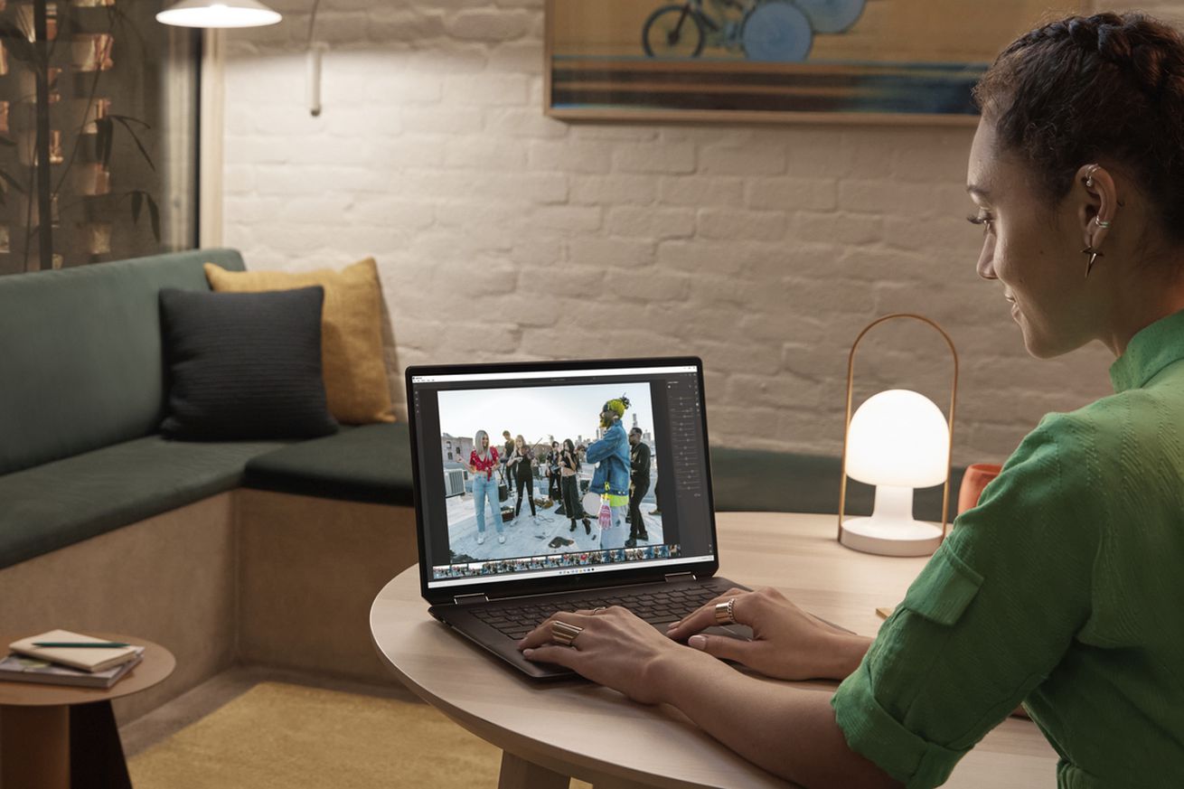 A user in a red dress types on the HP Spectre x360 16 in an evening room at a small wooden table. The screen displays a photo of a group of urban dancers in an outdoor scene in an editing program. To its left is a small lit lamp.