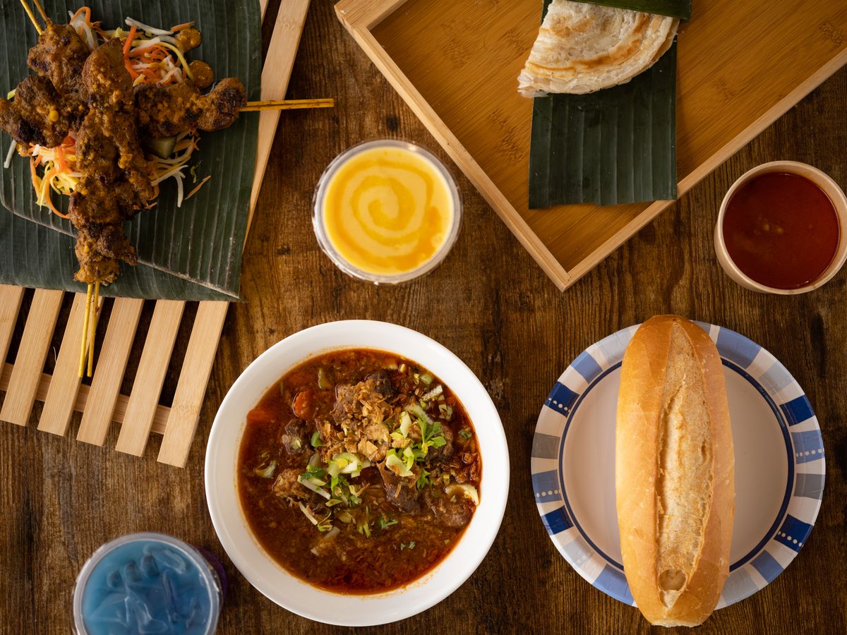 A spread of food from Salima Specialites, with chicken skewers on a banana leaf, thin-cut fried potato on a banana leaf, a bowl of dark red stew, a Vietnamese baguette on a white plate, and blue and mango-colored drinks in plastic cups on a wood surface.