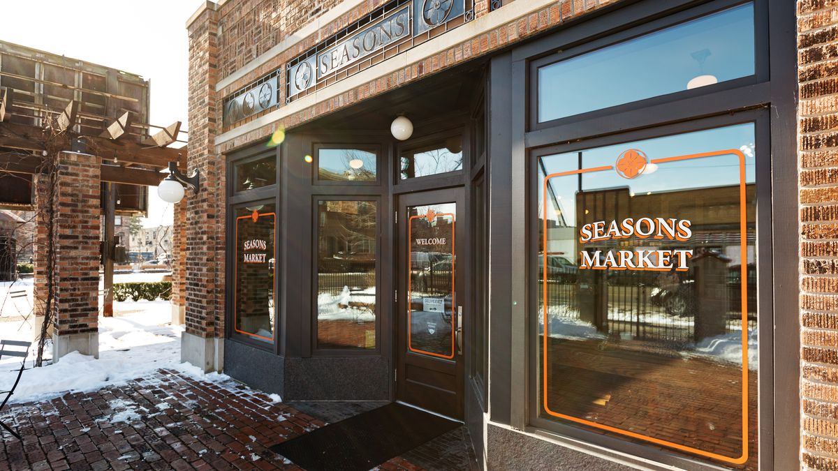 Photo of a brick building with glass windows with Seasons Market painted in white and orange on the right side window