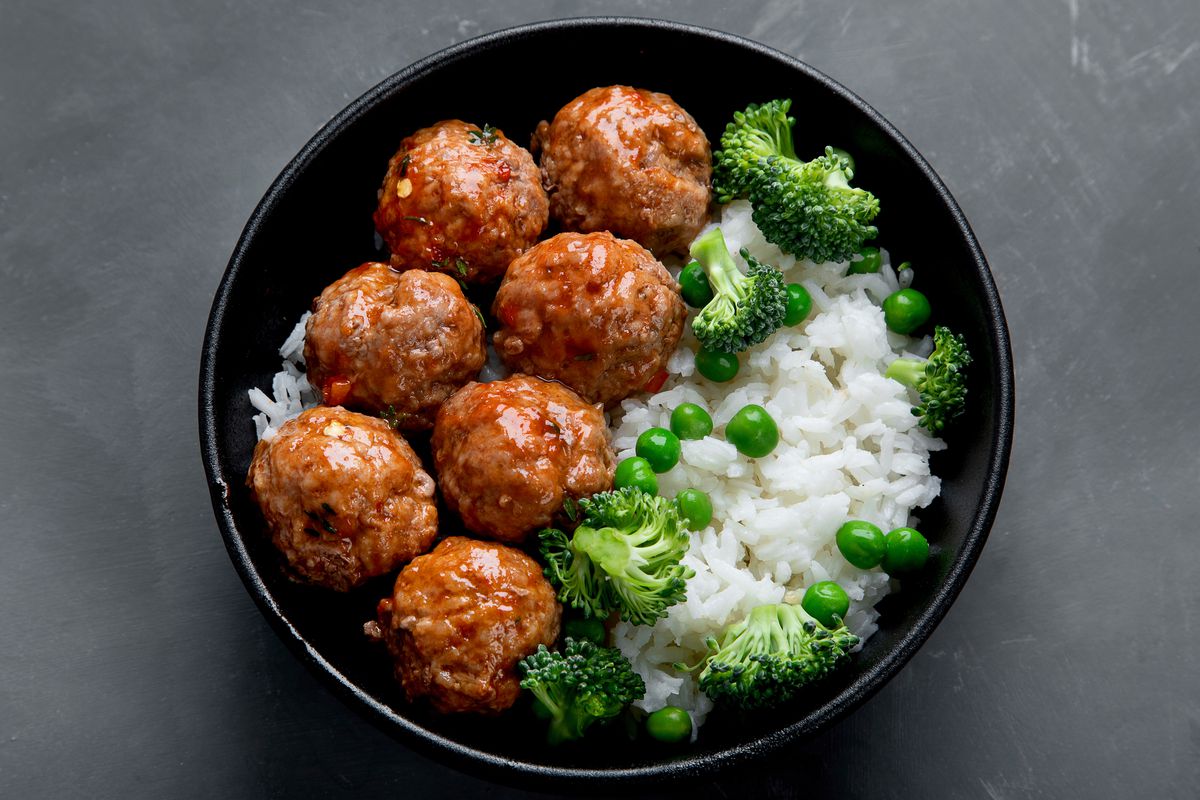 Seven meatballs in a black bowl sit alongside white rice, broccoli, and peas.