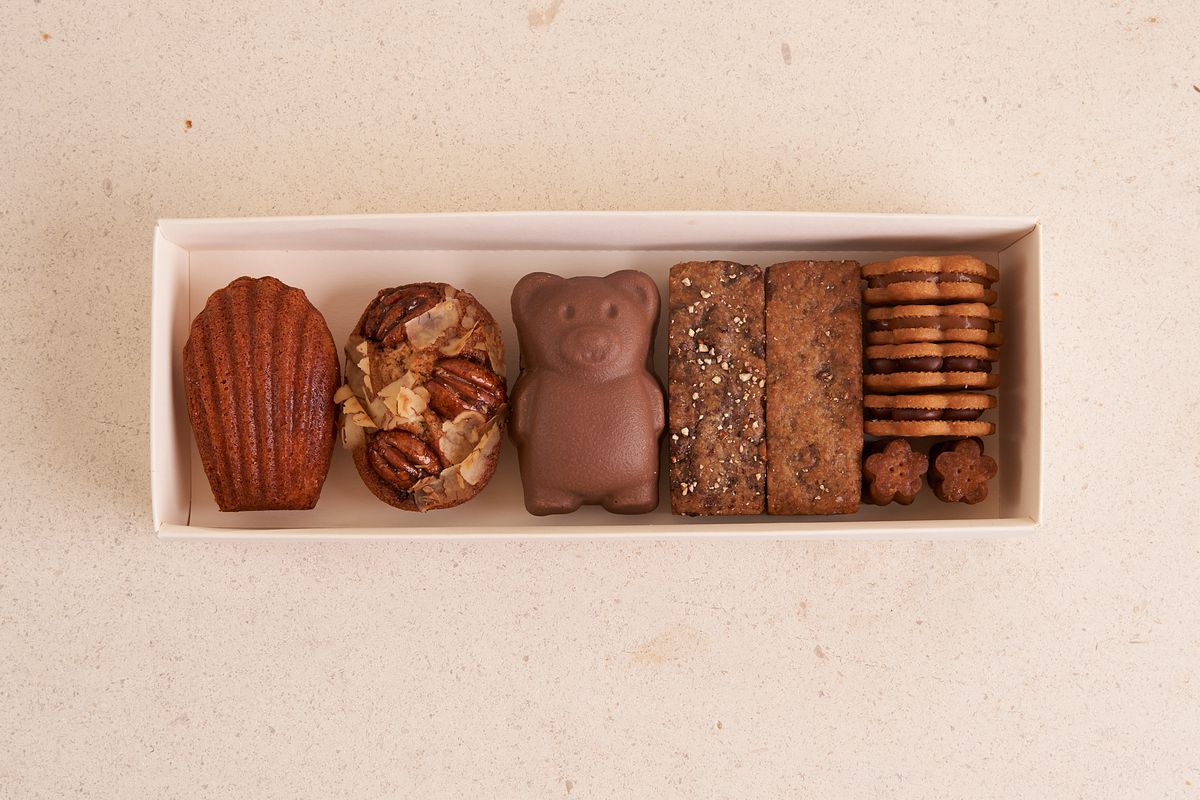 A white box sits on a white table and is filled with eight brown desserts including a madeleine, a bear-shaped treat, and cookies.