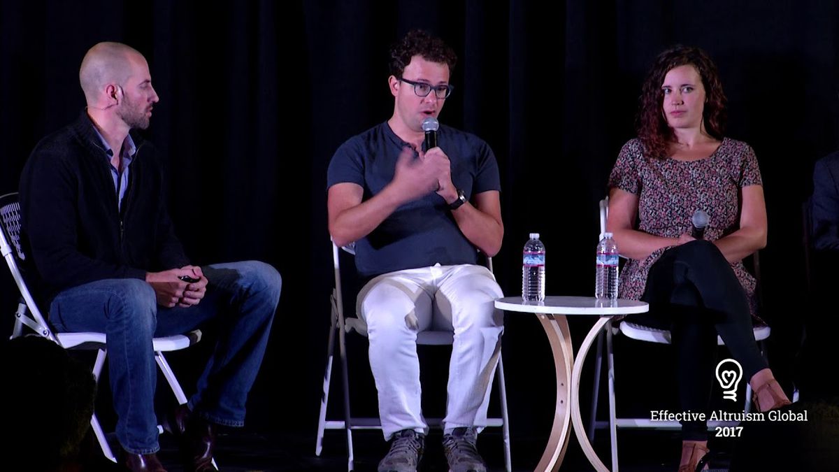 Anthropic CEO Dario Amodei holding a microphone during a panel discussion. On either side of him sit a man and a woman.