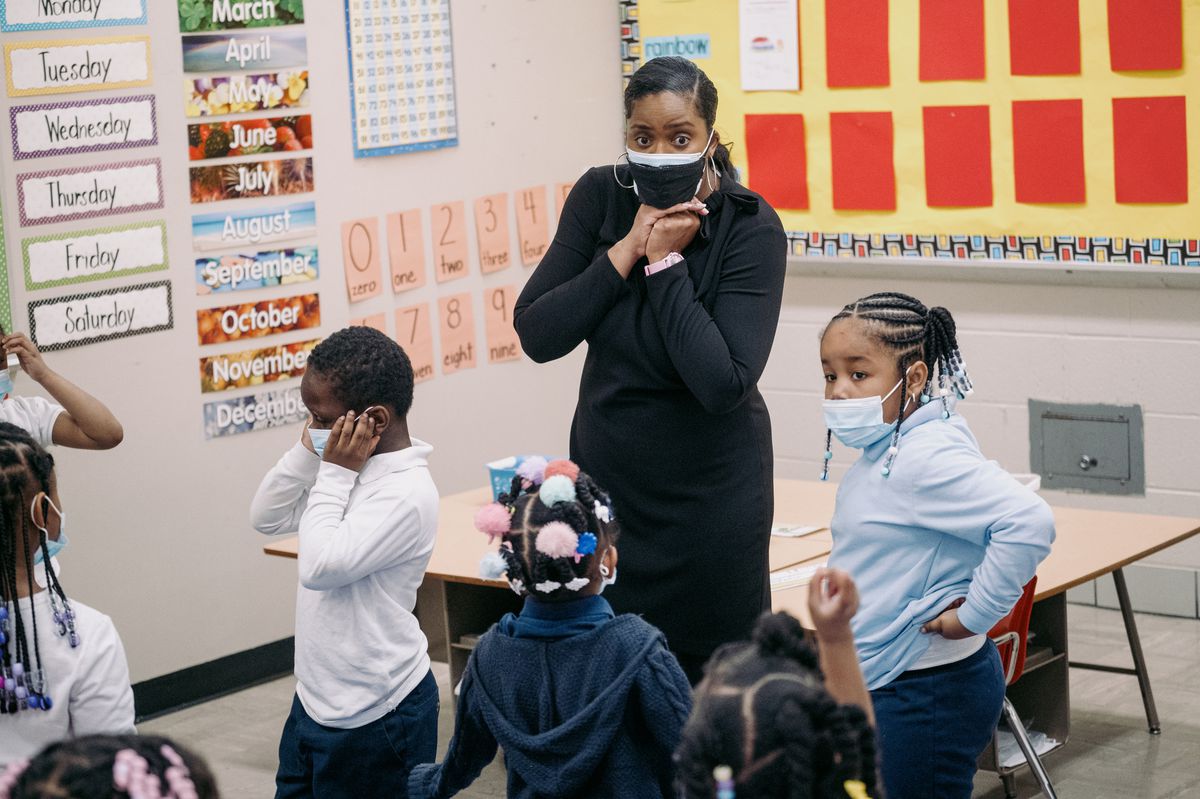 A woman teacher, wearing a black mask with a filtered protective mask underneath, looks on at her young students in the classroom as she grasps her hands underneath her chin.