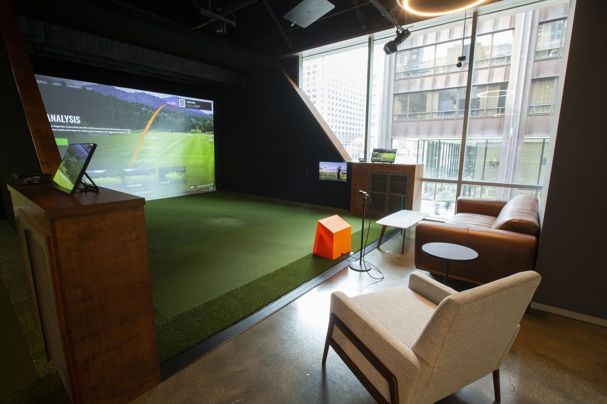 A golf simulator bay with a large screen and putting green before two lounge chairs.