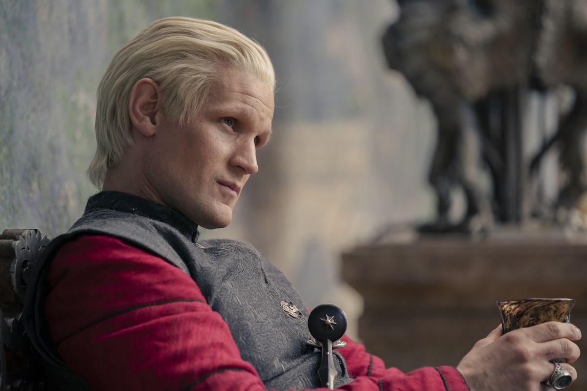Daemon Targaryen, greased, wearing a red shirt and gray cape, glares at someone as he holds his wine glass in House of the Dragon