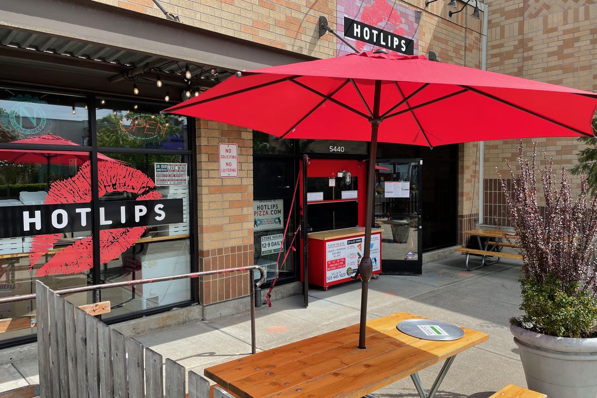 The exterior of a pizza shop emblazoned with a lipstick print logo on the window. A picnic table with a large red umbrella covering it sits in front.