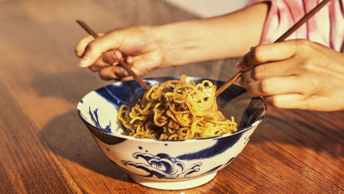 Ramen noodles in a blue and white ceramic bowl with a person lifting the noodles with chopsticks.