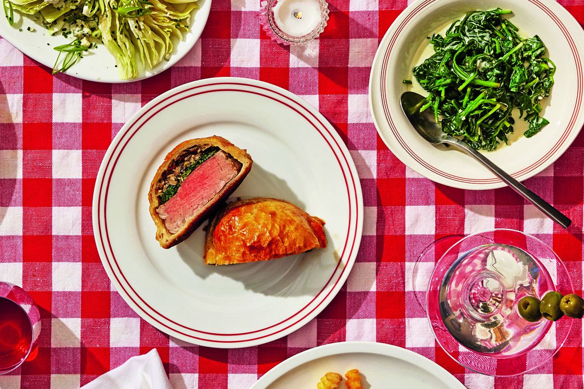 Beef Wellington on a plate cut in half with cross-section of steak and filling showing. Plate is on a checkered tablecloth alongside cooked spinach.