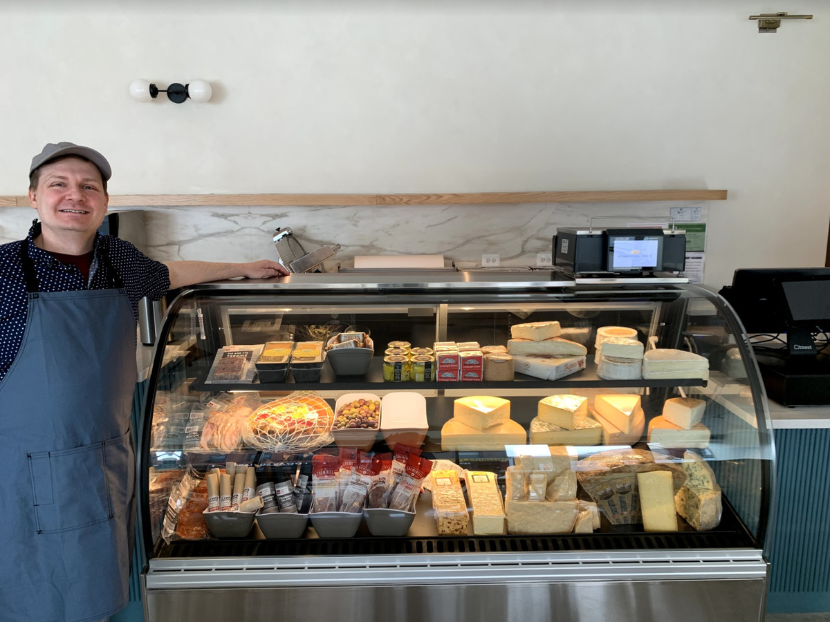 A white man stands next to a glass display case filled with cheese.