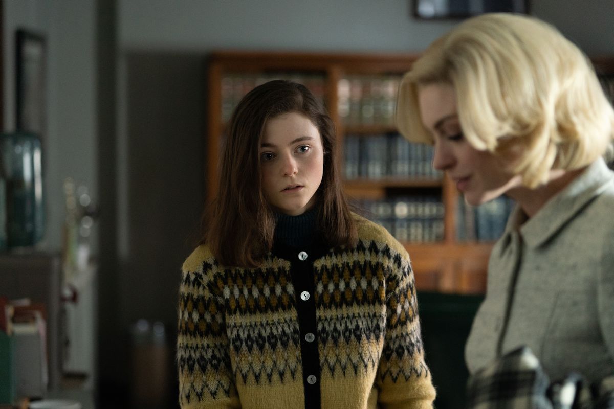 Thomasin McKenzie stands in a kind of ugly brown sweater looking on nervously at a blonde Anne Hathaway doing office work, glamorously, in the movie Eileen
