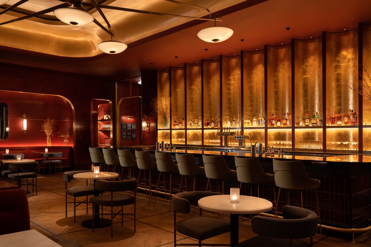 A long bar with shimmering golden alcoves behind it and a row of bar stools. Small tables are in the foreground. 
