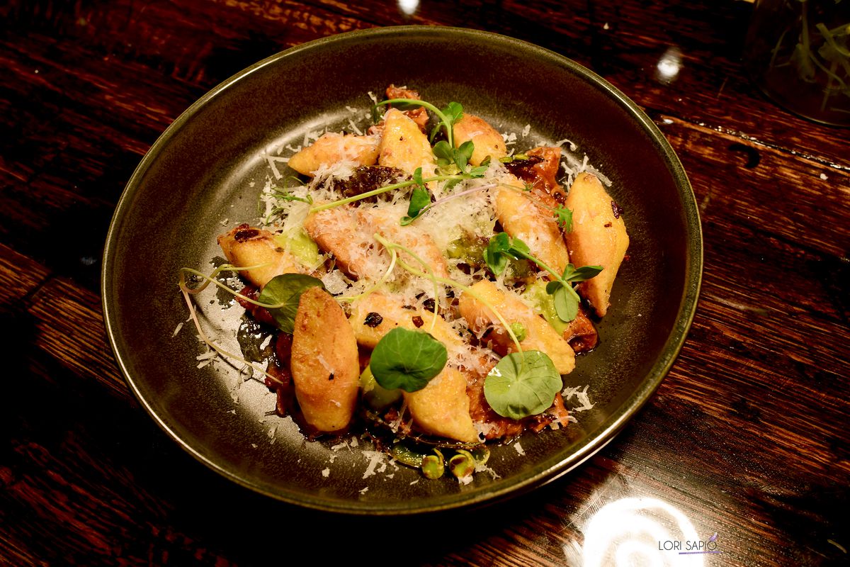 A small pile of orange-colored yucca dumplings sit on a charcoal-colored plate and are topped with shredded cheese and herb leafs.