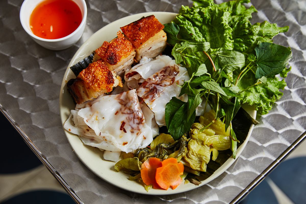 A white plate with cubes of crispy pork belly, steamed rice rolls, leafy green lettuce, and other vegetables set on a stainless steel table with a small white cup of orange dipping sauce placed nearby