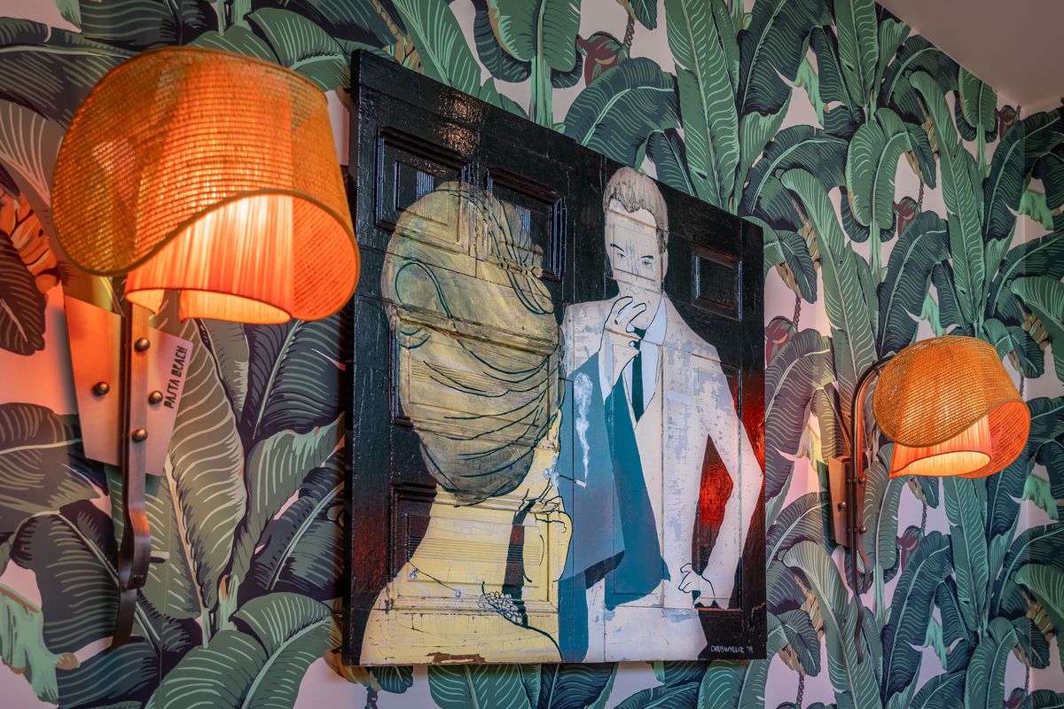 A painting of a man and woman in formal attire hangs on a restaurant wall that is covered with banana leaf wallpaper.