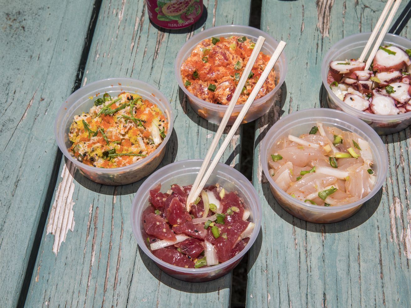 Various kinds of poke in plastic containers with drinks and chopsticks on a light blue picnic table.