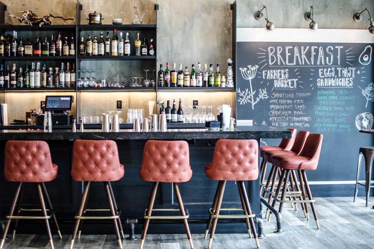 Pale red cushioned barstools line a black bar behind which stands a wall and shelving for liquor bottles