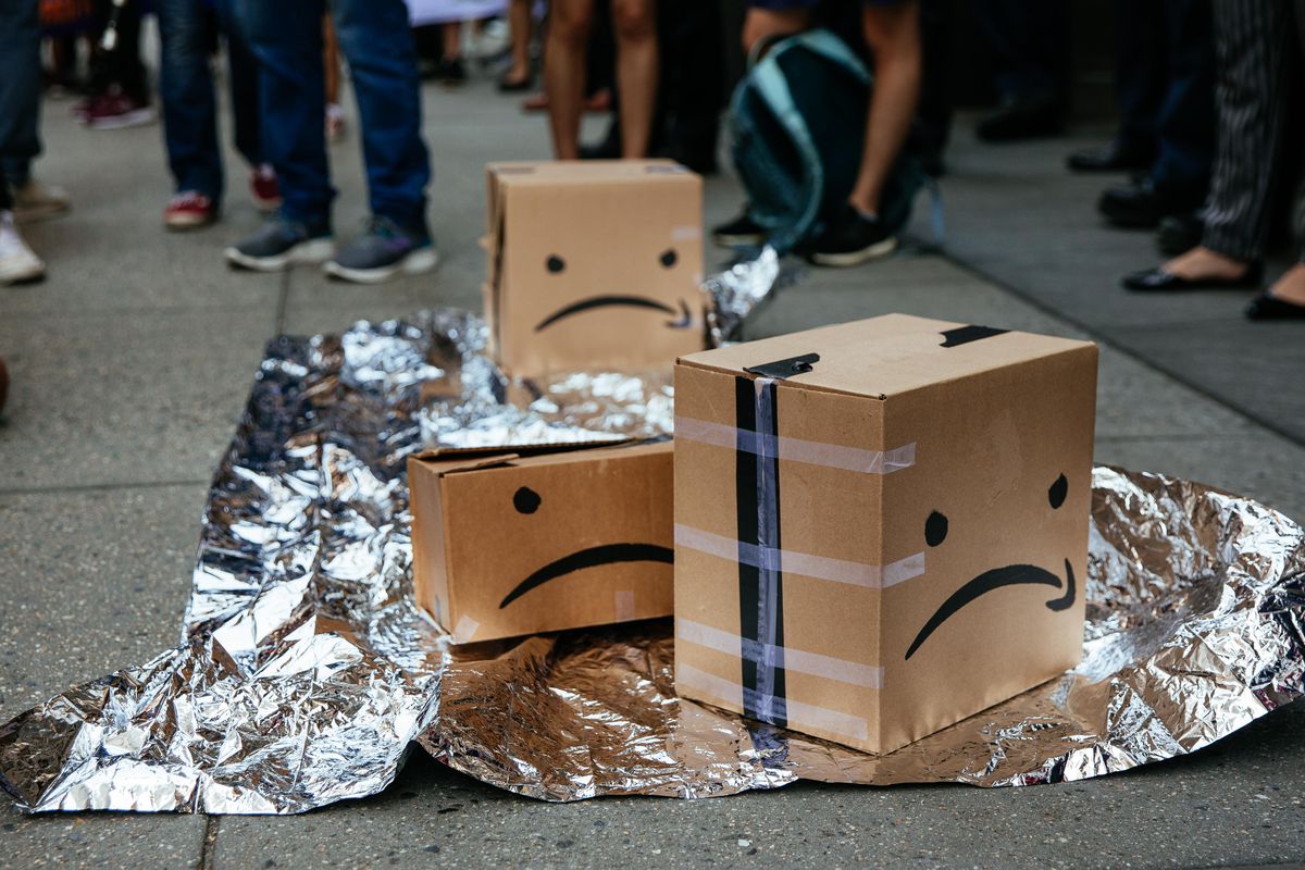 Amazon boxes with drawn-on sad faces (the Amazon logo upside down) sit on the pavement atop a silver groundsheet.