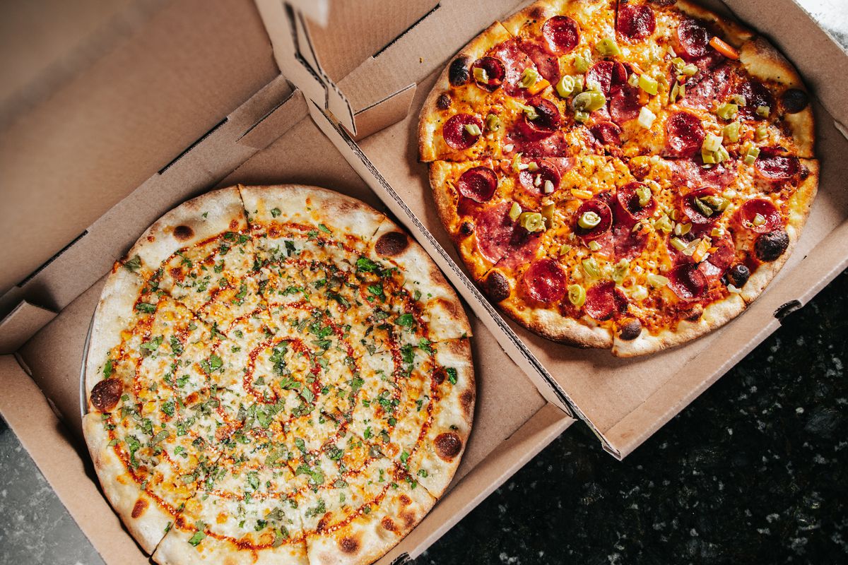 Two pizzas, both served in carboard takeout boxes. On the left is an elote pie, on the right is a pepperoni pizza 
