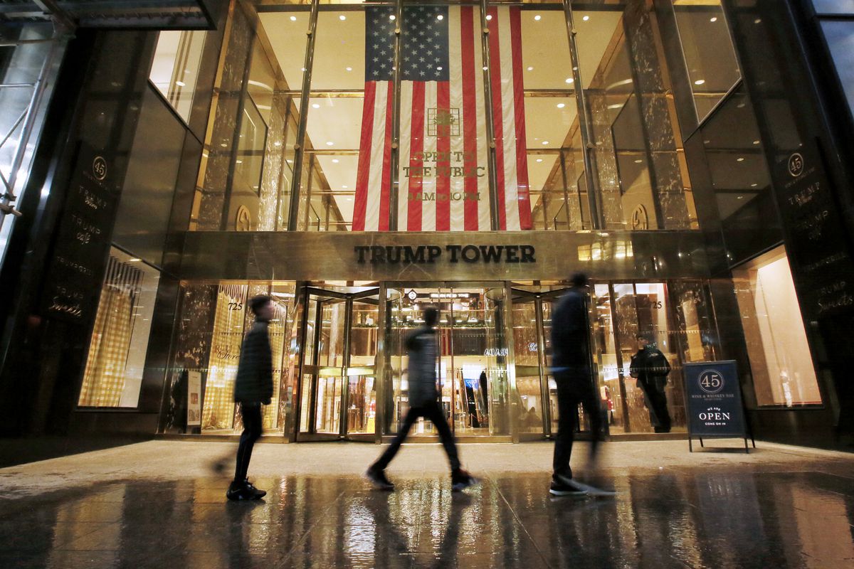 In the fading light, the golden facade of the tower’s ground floor glitters, black letters spelling out “Trump Tower” over glass revolving doors. Above the sign are large windows with golden panes; through them a massive US flag is visible. Three figures, blurs dressed in black, walk past the entrance.