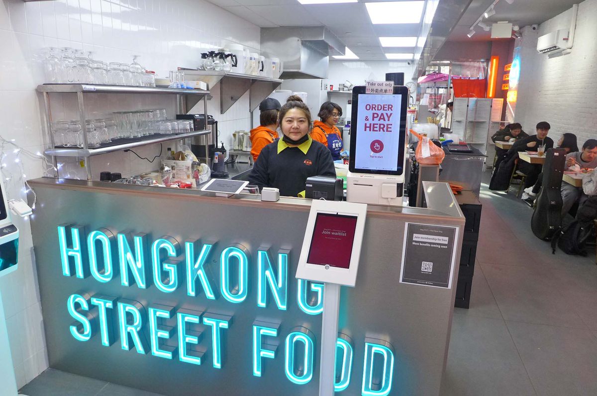 A woman stands behind a counter with a neon sign that reads Hong Kong Street Food.