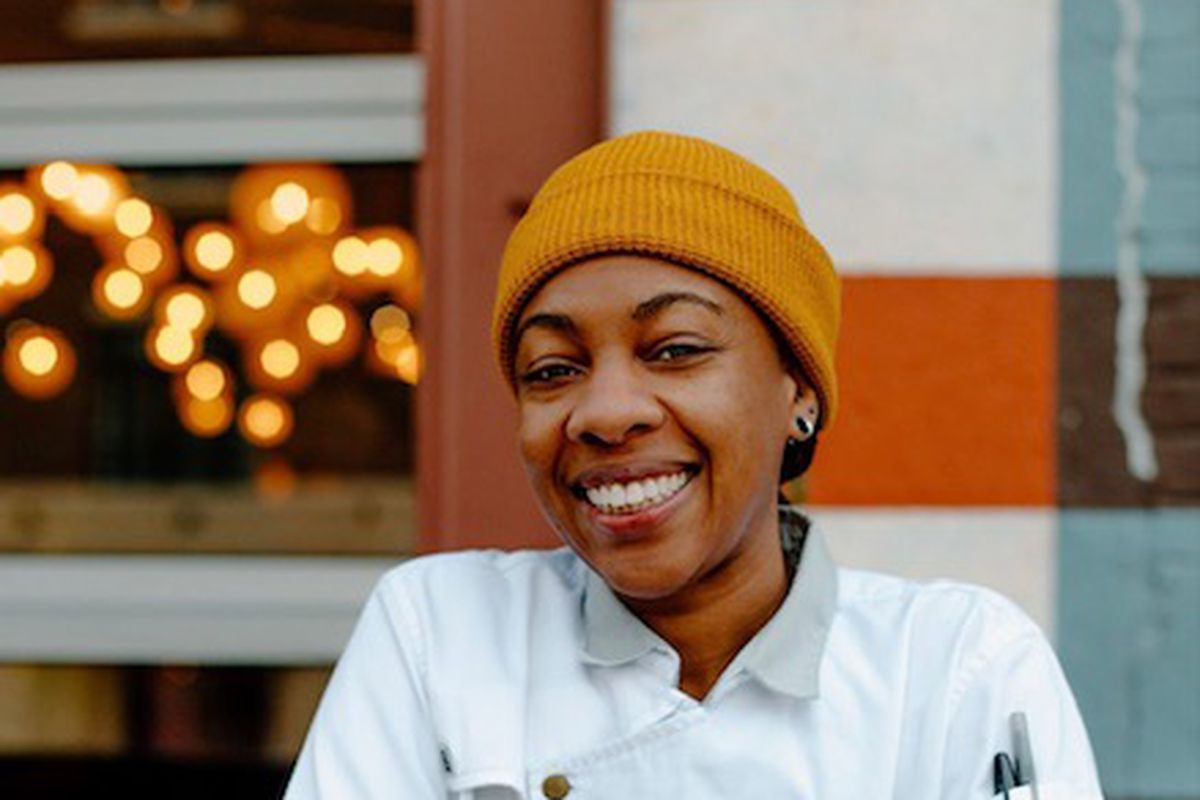 Chef Ashleigh Shanti in chef’s whites and a beanie, standing against an outdoor railing.