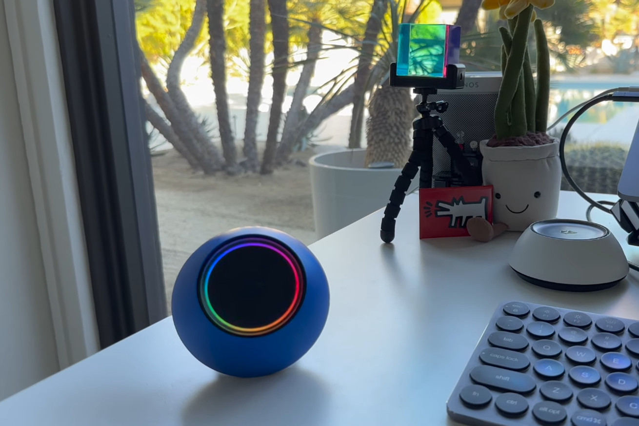 Screenshot from a Josh.ai YouTube video showing a blue smart speaker with an RGB ring around it. The speaker is on a white desk; a Palm Springs courtyard is visible behind.