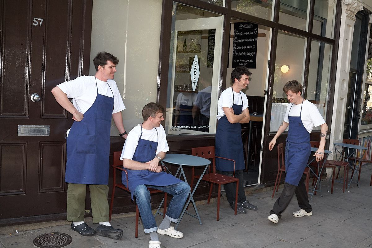 Four men in white t-shirts and blue aprons stand outside a wine bar, standing together around blue tables.