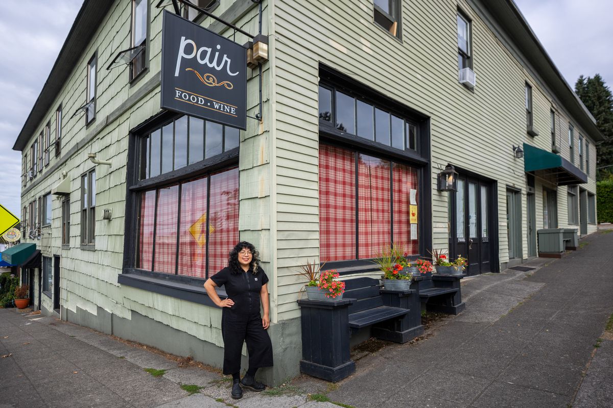 A Latina woman in glasses and a dark jumpsuit stands outside a storefront that reads ‘Pair.”