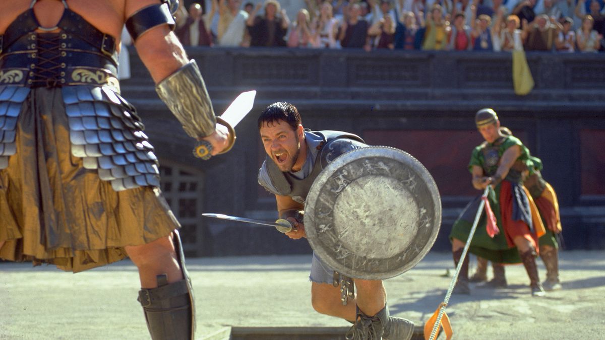 Russell Crowe as General Maximus Decimus Meridius holding a sword and shield and lunging at a gladiator in Gladiator.