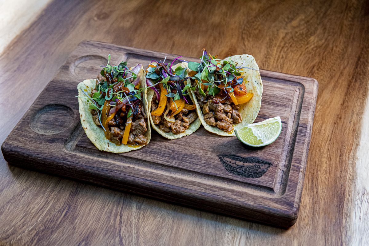 Three neatly plated al pastor tacos atop a wooden carving board from D Boca N Boca in Atlanta.