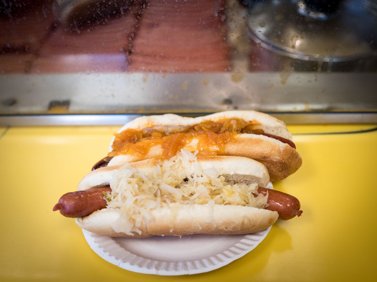 Two hot dogs sitting side by side on a white paper plate placed on a yellow tables. One of the hot dogs is topped with sauerkraut and another is topped with an orange sauce.