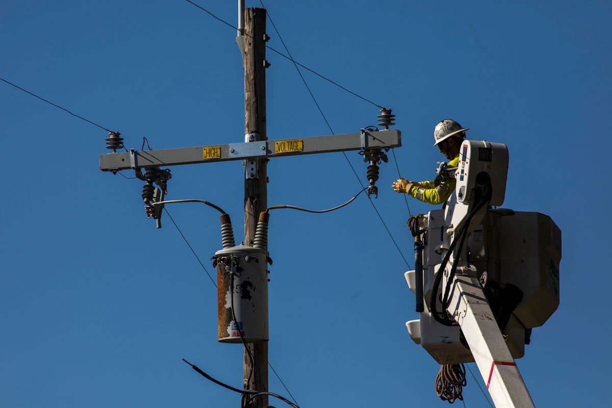 A worker in a hardhat and yellow vest using a crane to reach power lines during an inspection.