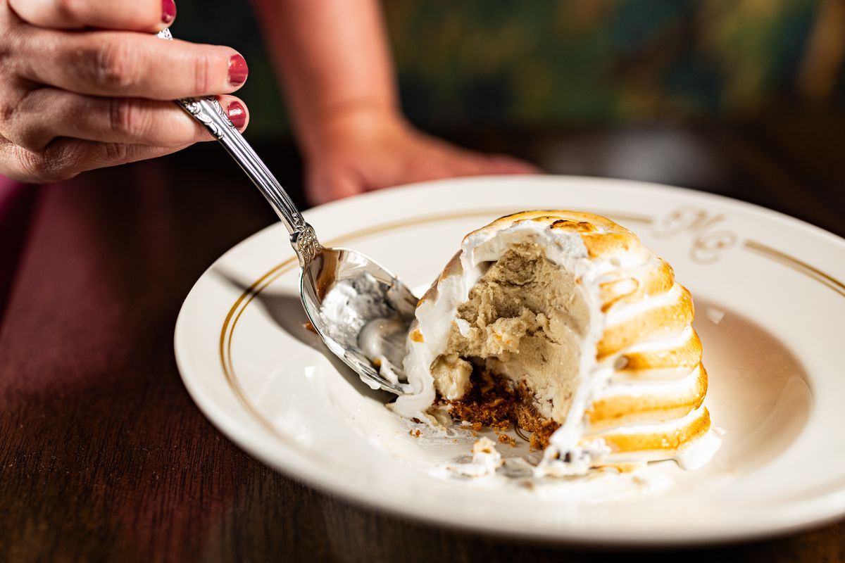 A woman’s hand holds a large spoon that scoops into half of a Baked Alaska, which is banana pudding inside.