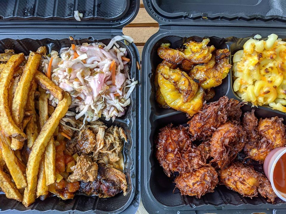 Overhead view of two black takeout containers on a picnic table. One contains jerk chicken with sides of fries and a slaw, while the other contains fried, coconut-encrusted shrimp with mac and cheese, plantains, and a small plastic cup of dipping sauce.