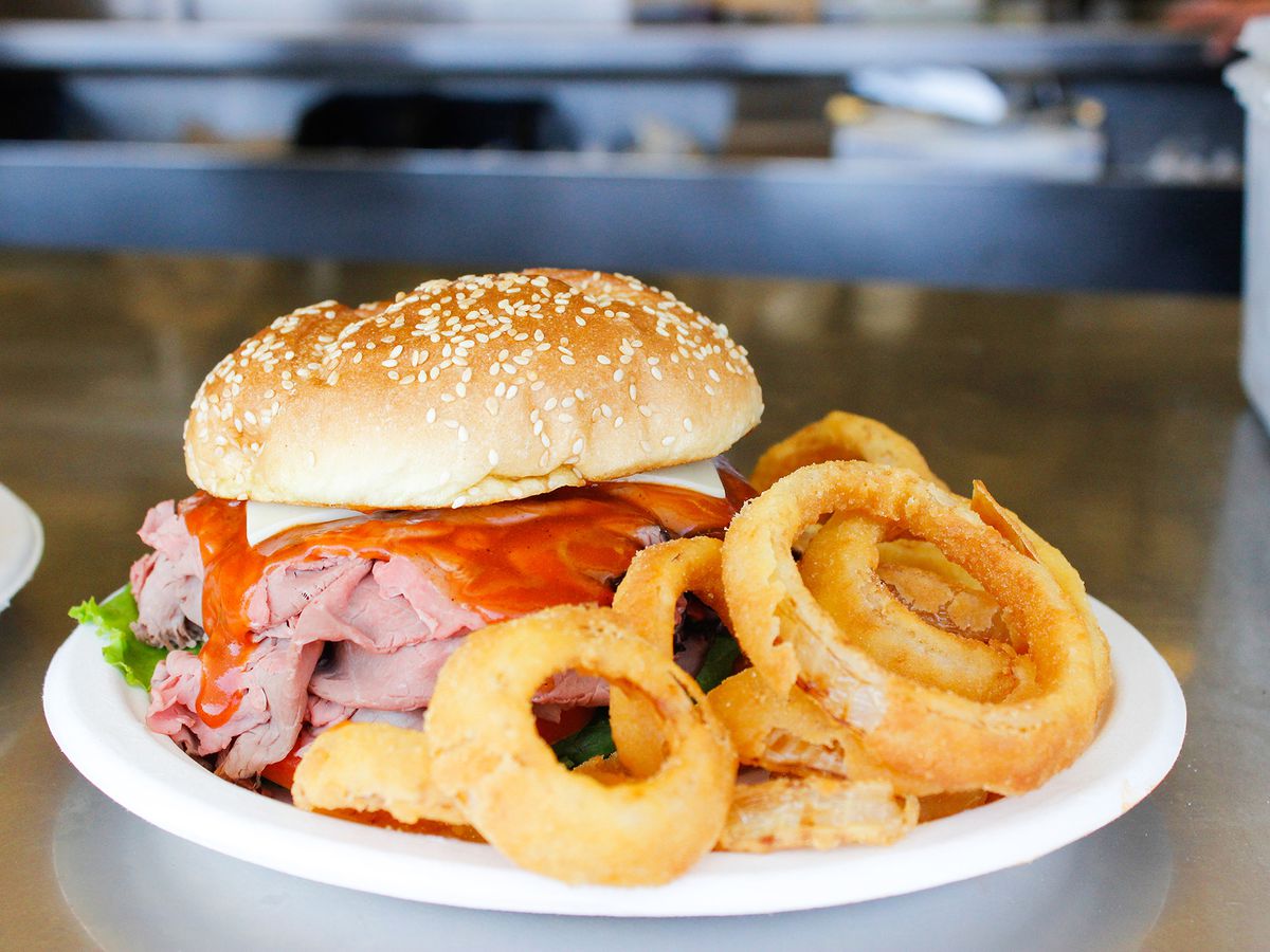 A roast beef sandwich with cheese, mayo, and barbecue sauce sits on a paper plate, accompanied by thick onion rings.