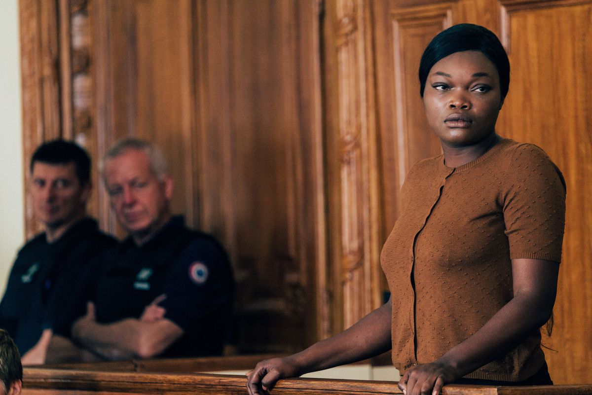Guslagie Malanda stands on trial as two officers look on in Saint Omer. Her brown-orange top matches the wood of the court.