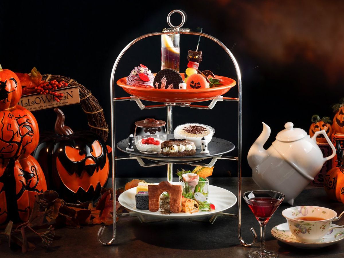 An afternoon tea set with three-tiered tray with Halloween cookies and candies, and finger sandwiches. White teapot set and pumpkins surround it.