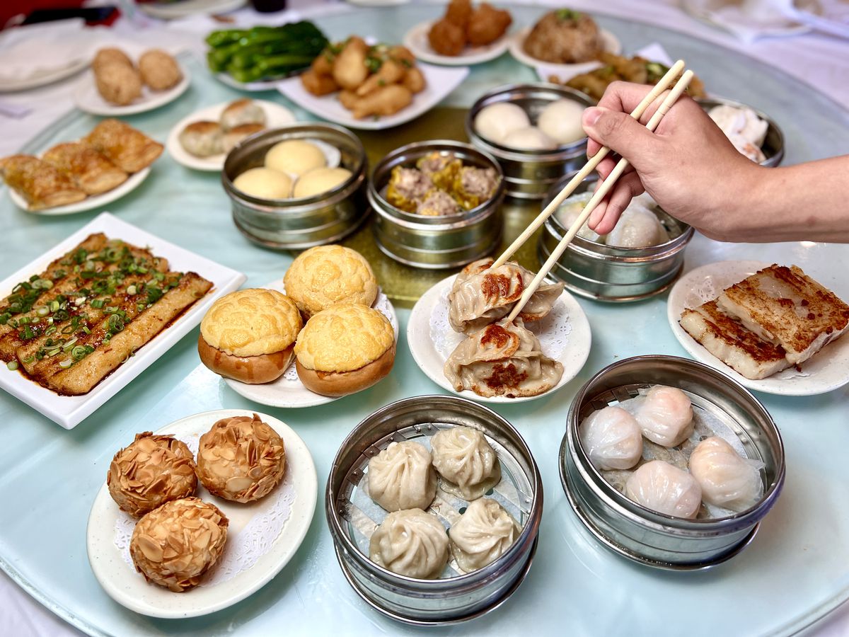 a hand with chopsticks picks up a dumpling from a tray, atop a table at Golden Dim Sum that’s filled with dumplings, bread, and fried goodies.