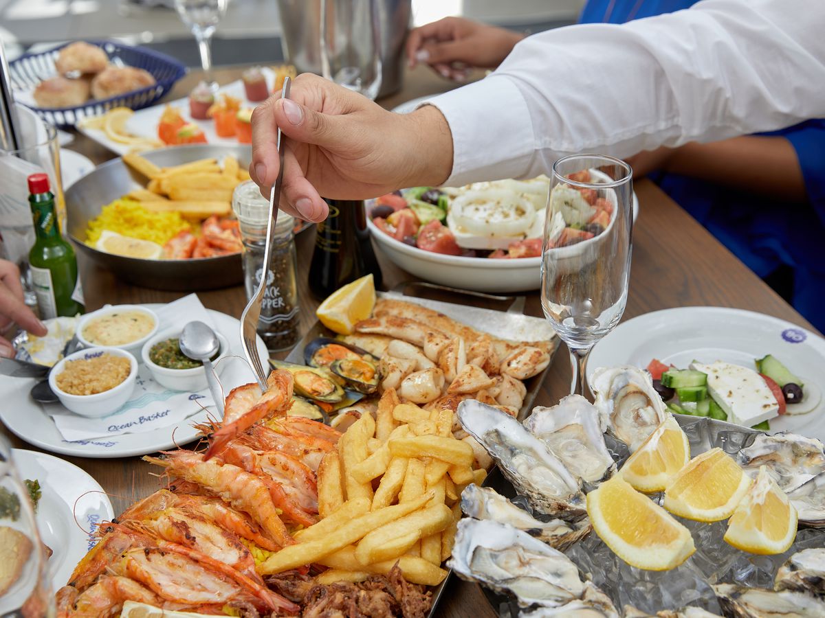 A hand reaches across a table with a fork to take an item from a large platter of various seafood, on a table filled with other dishes including a tray of oysters, fried seafood with French fries, Greek salad, and glasses for wine
